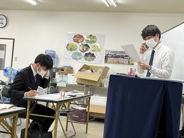 スーツとマスクを着用した2人がデスクに座っている様子