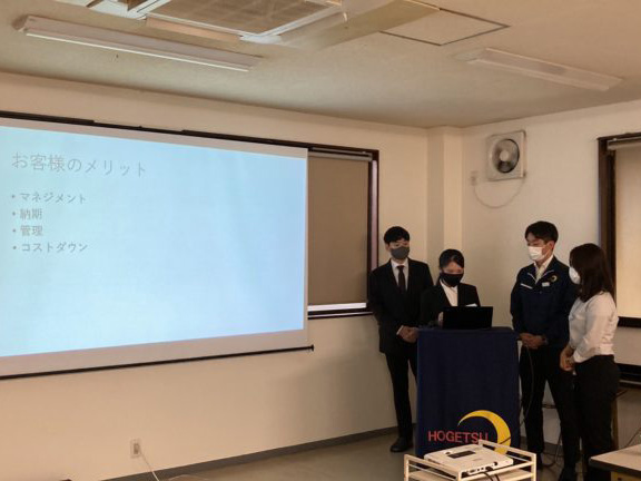 緑の作業着を着た女性が手に棒を持ってプレゼンテーションをしている